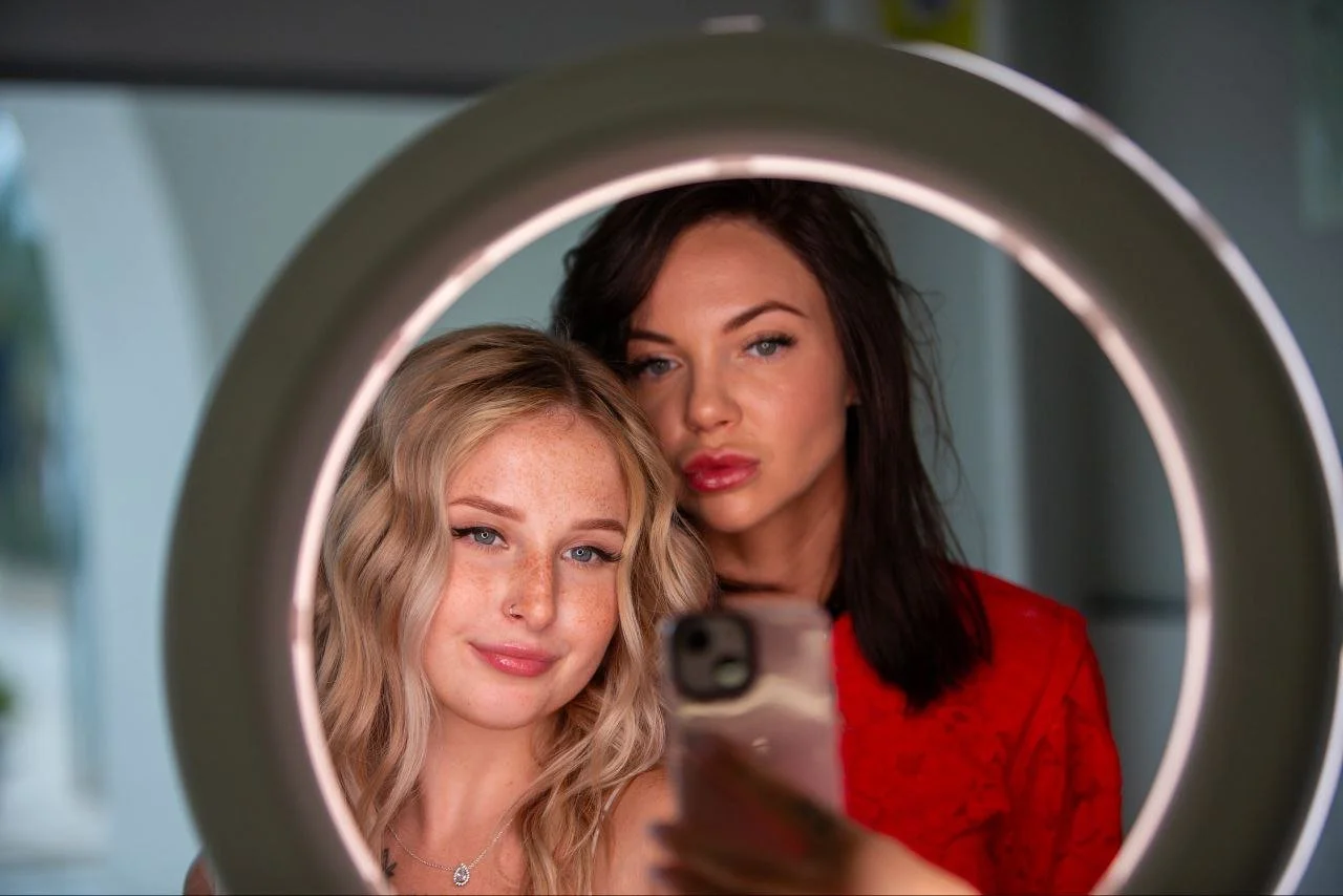 Two women taking a selfie behind a ring light with a smartphone, one with blonde wavy hair and the other with dark straight hair, both looking at the camera.