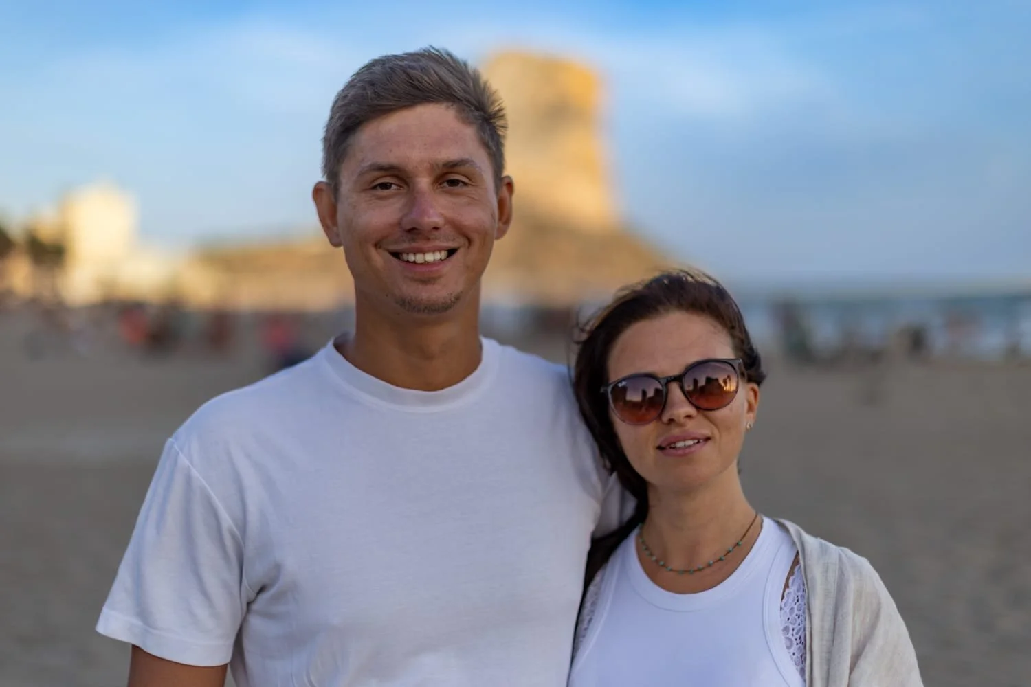 A smiling man and woman stand on a beach with a large rock formation in the background. The man is wearing a white t-shirt and the woman is wearing sunglasses and a white top.