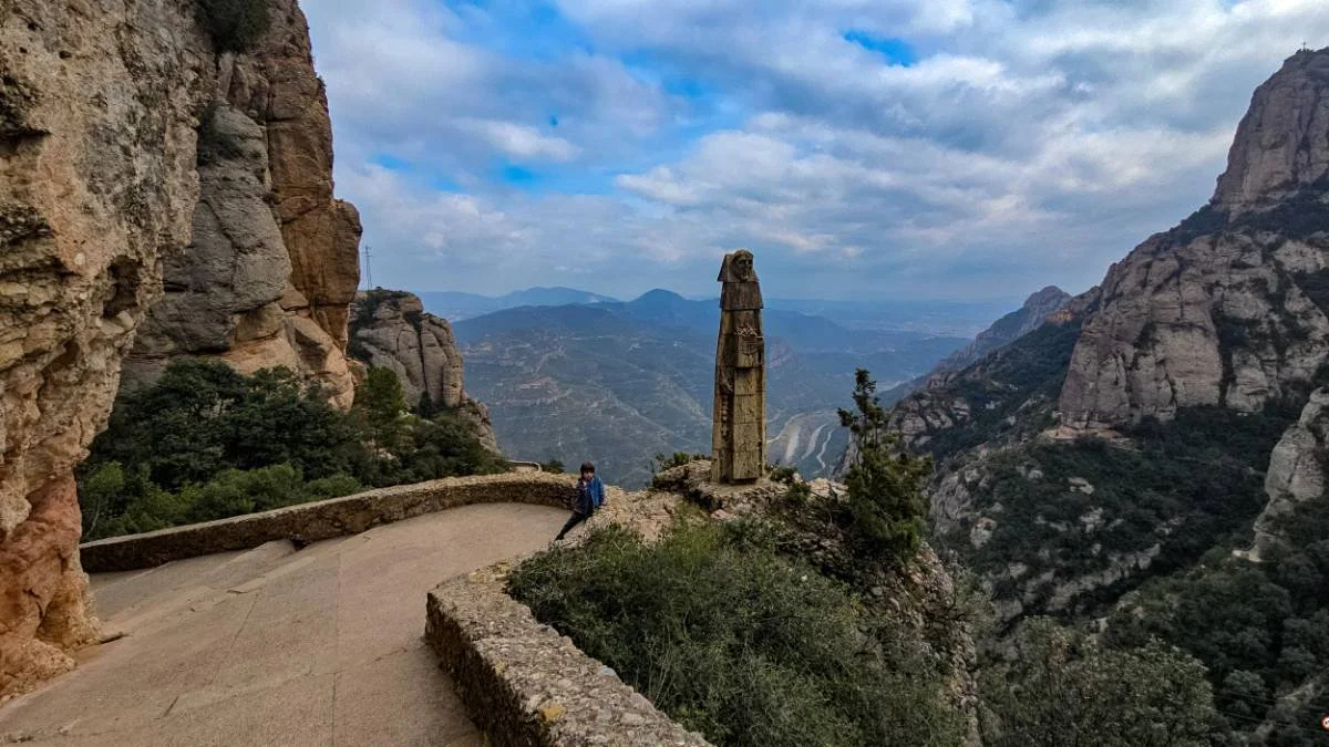 A person sitting on a stone pathway with a mountainous landscape in the background, including cliffs, trees, and a stone monument or sculpture.
