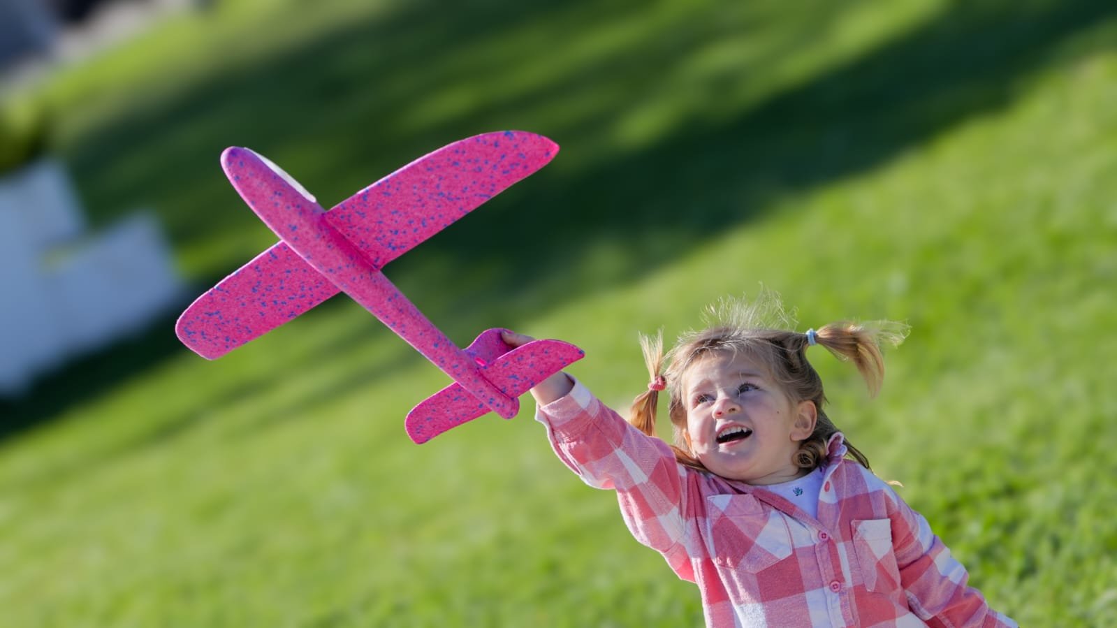 Young girl holding a pink foam toy airplane and looking up outdoors on a sunny day.