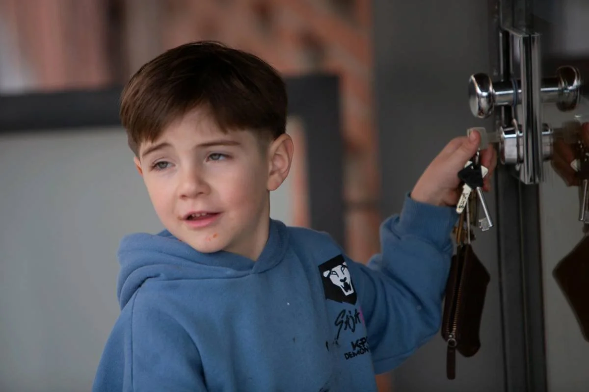 A young boy with brown hair, wearing a blue hoodie, is turning a set of keys in a lock on a locker.