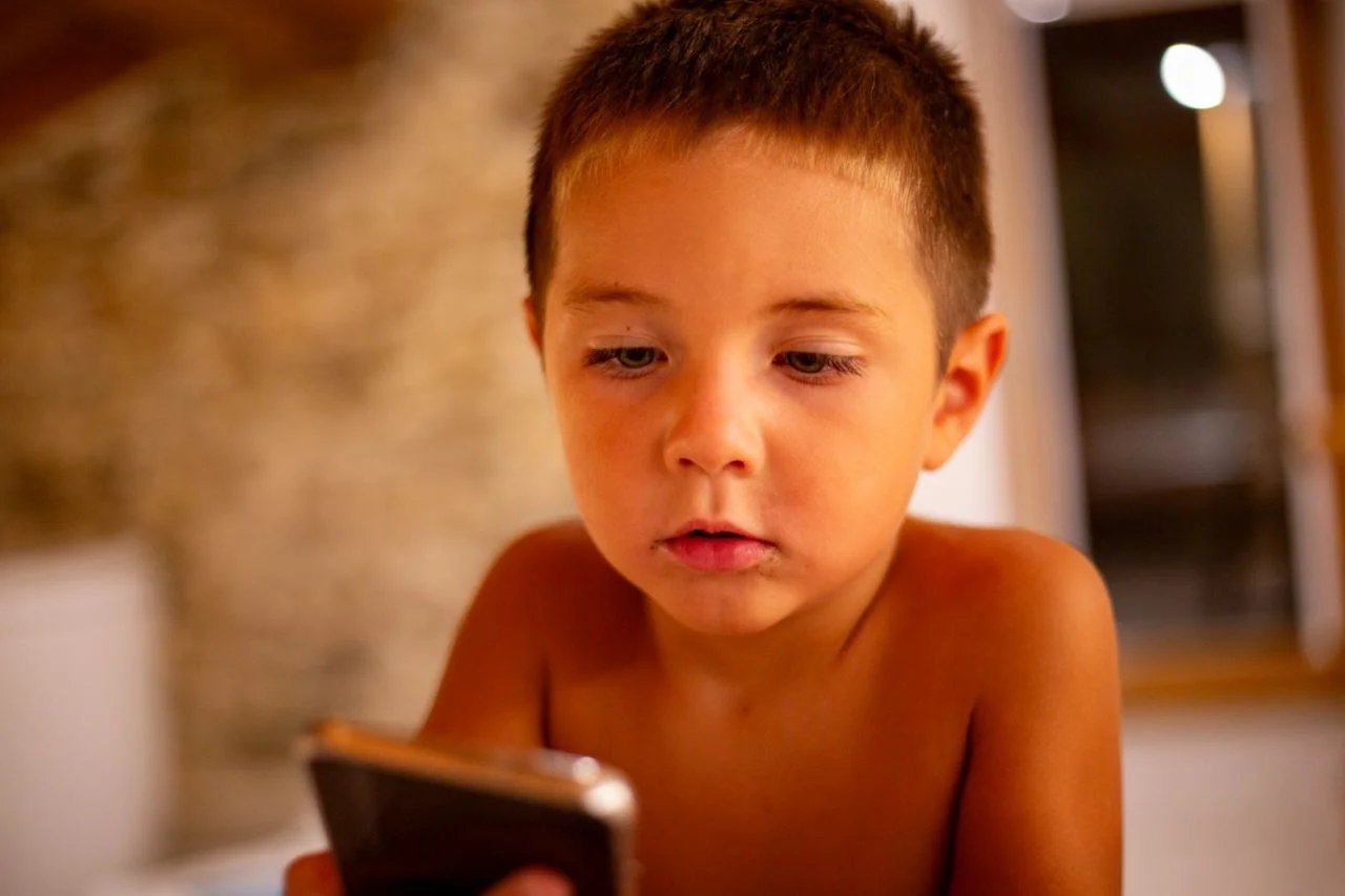 A young boy with short brown hair looking at a smartphone, indoors with a blurred background.