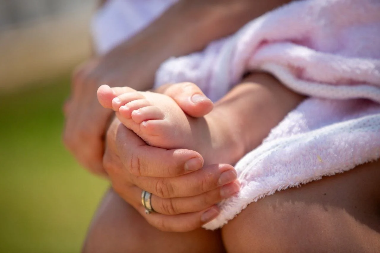 Close-up of an adult holding a baby's foot with a towel wrapped around the baby's body, outdoors with a blurred grassy background.