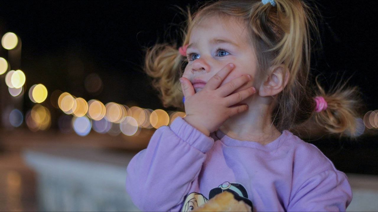 A young girl with blonde hair tied in pigtails, wearing a purple shirt, looks thoughtful with her hand on her face at night, with blurred city lights in the background.