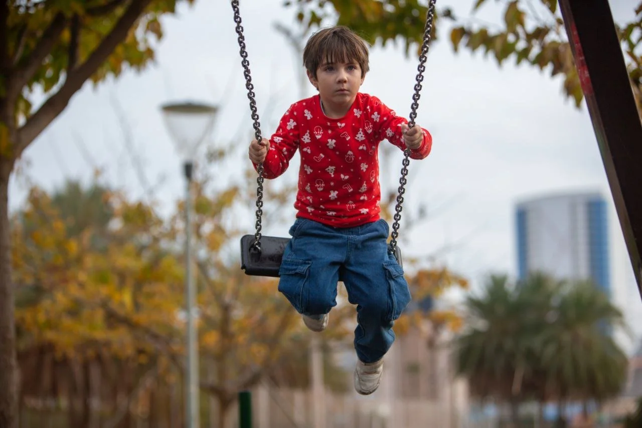 Child in a red long-sleeve shirt with white patterns, blue pants, and sneakers swinging on a playground swing set.