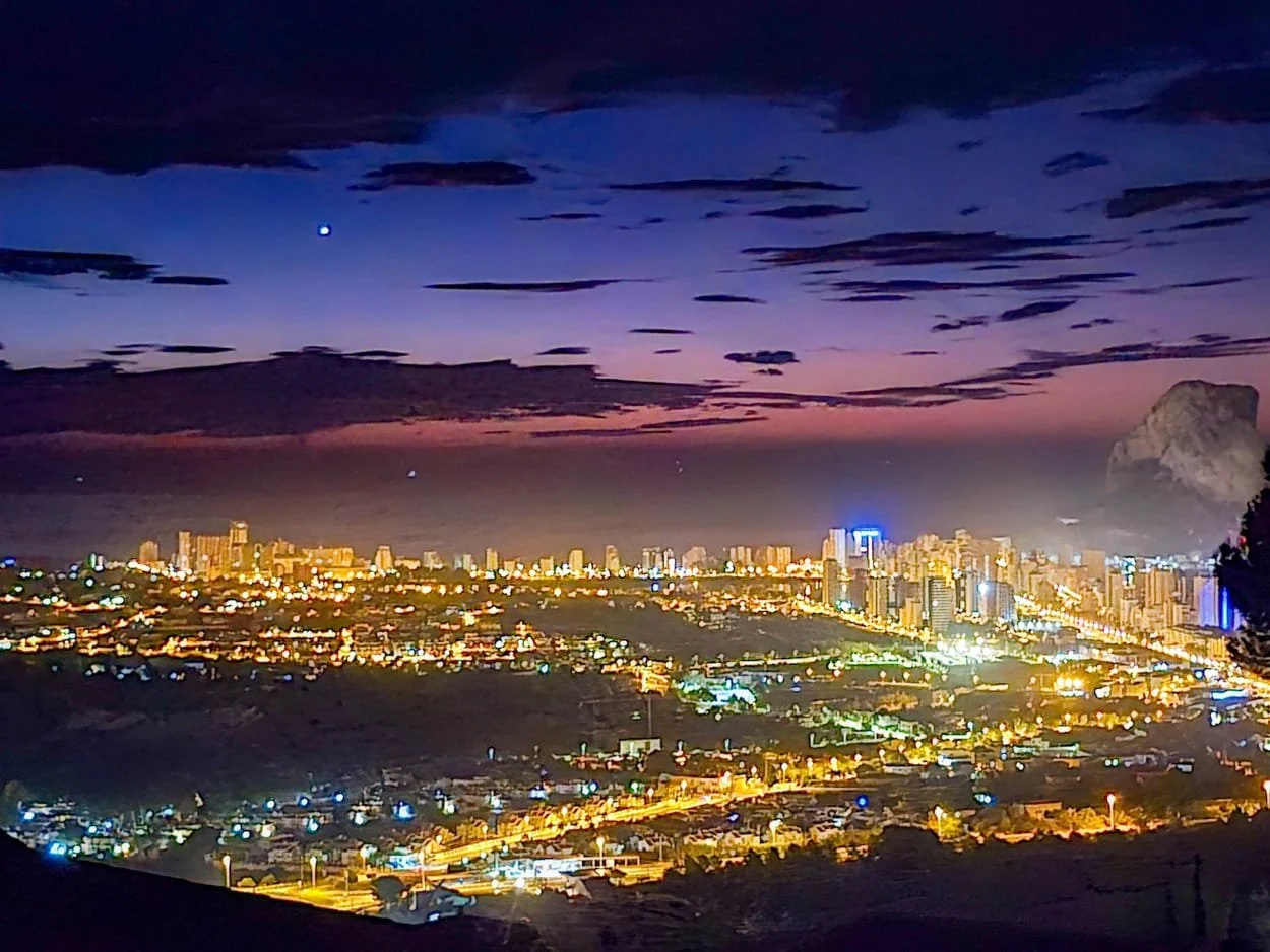 Nighttime cityscape with illuminated buildings and streets, a dark sky with some clouds, a visible moon, and large rocky formation on the right side.