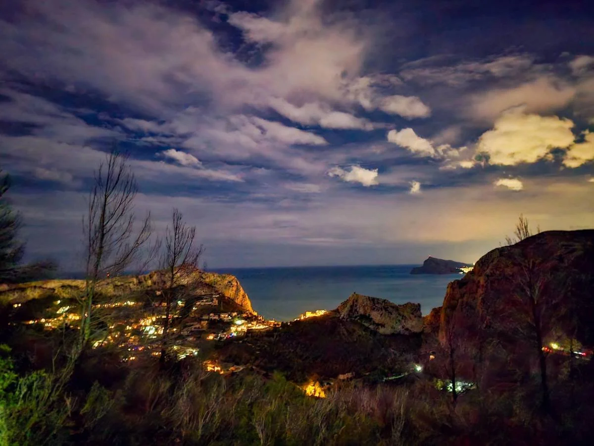 Nighttime coastal scene with illuminated town, rocky hills, and cloudy sky over the ocean.