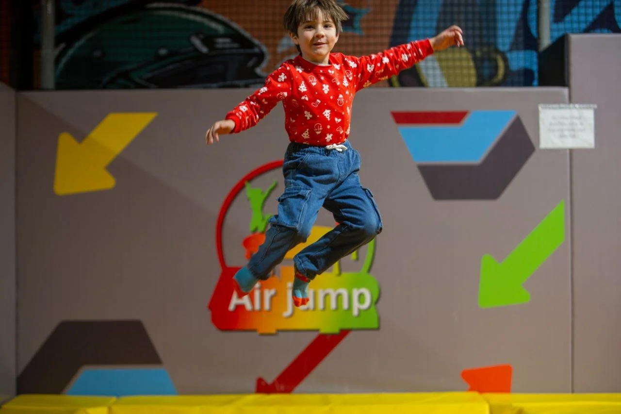 A young boy wearing a red sweater with holiday patterns and blue jeans jumping on an indoor trampoline at an air jump facility.