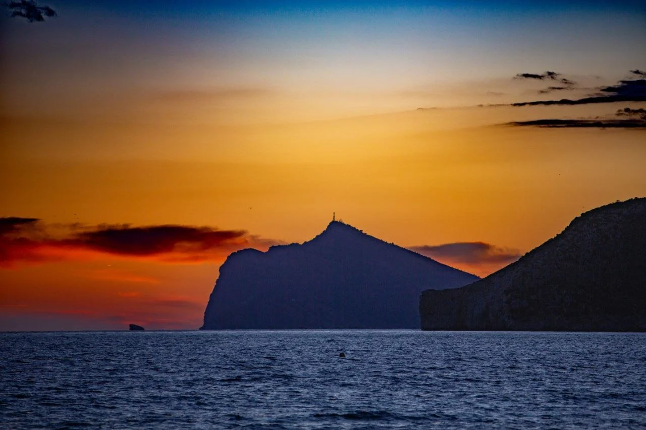 Sunset over a body of water with silhouetted mountains, including a prominent peak with a small structure or antenna on top, and a colorful sky with clouds.