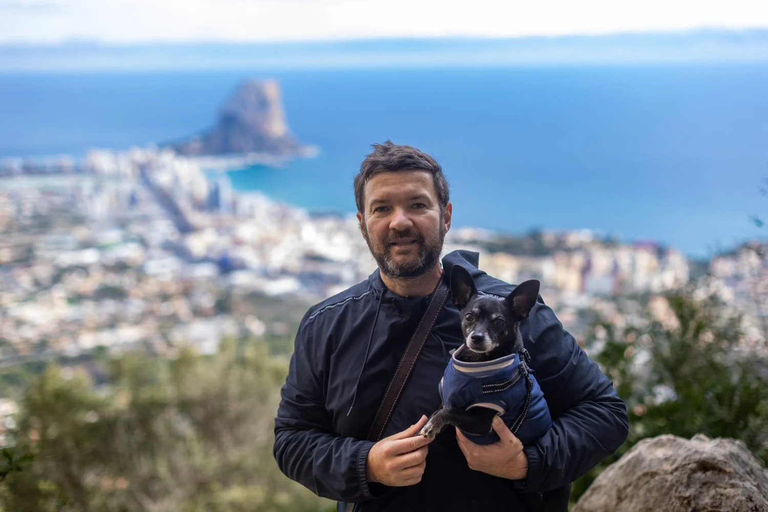 A man with a beard holding a small black dog wearing a blue jacket, with a blurred cityscape and ocean in the background.