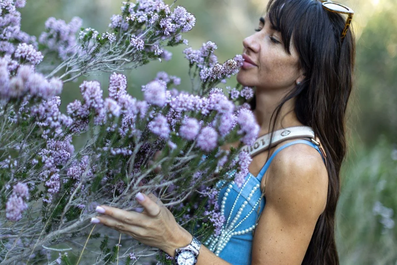 A woman with long dark hair and freckles is smelling purple wildflowers outdoors, wearing a blue dress with pearl accents, sunglasses on her head, and a wristwatch.