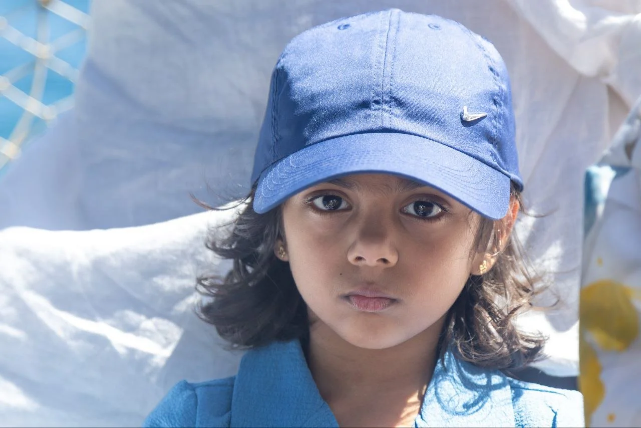 A young girl with dark hair wearing a blue Nike cap and blue shirt, looking directly at the camera.