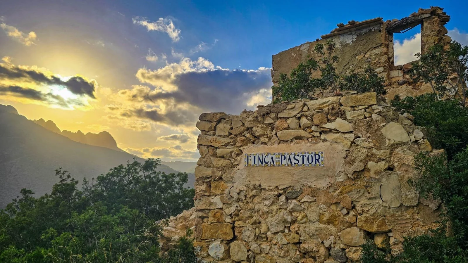 Sunset over mountains with ruins of a stone building and a sign that reads 'Finca Pastor', surrounded by green vegetation.