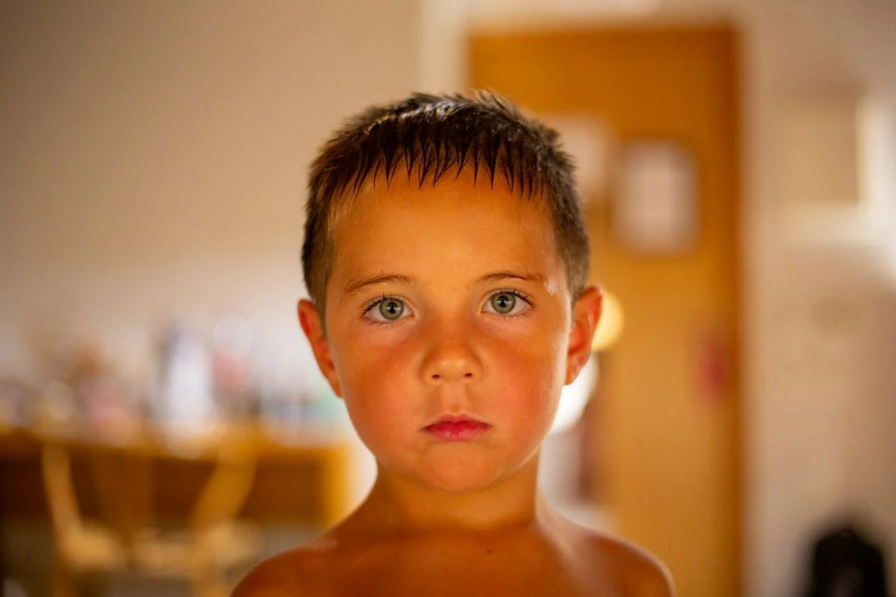 A young boy with wet hair and a serious expression standing in a warmly lit indoor space.