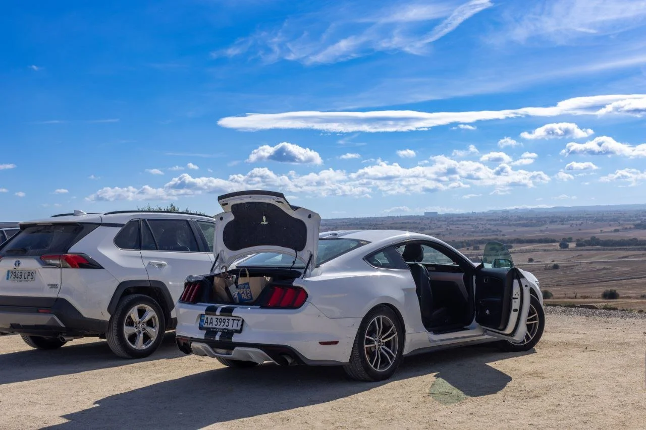 A white sports car with open doors and trunk parked on a dirt area next to a gray SUV, with a landscape and blue sky with clouds in the background.