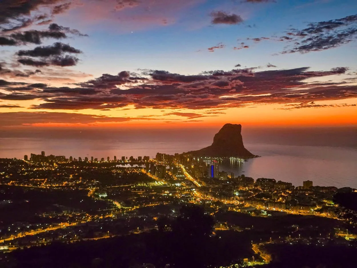 A cityscape at sunset near the ocean with a large rock formation or mountain in the water, and city lights beginning to twinkle.