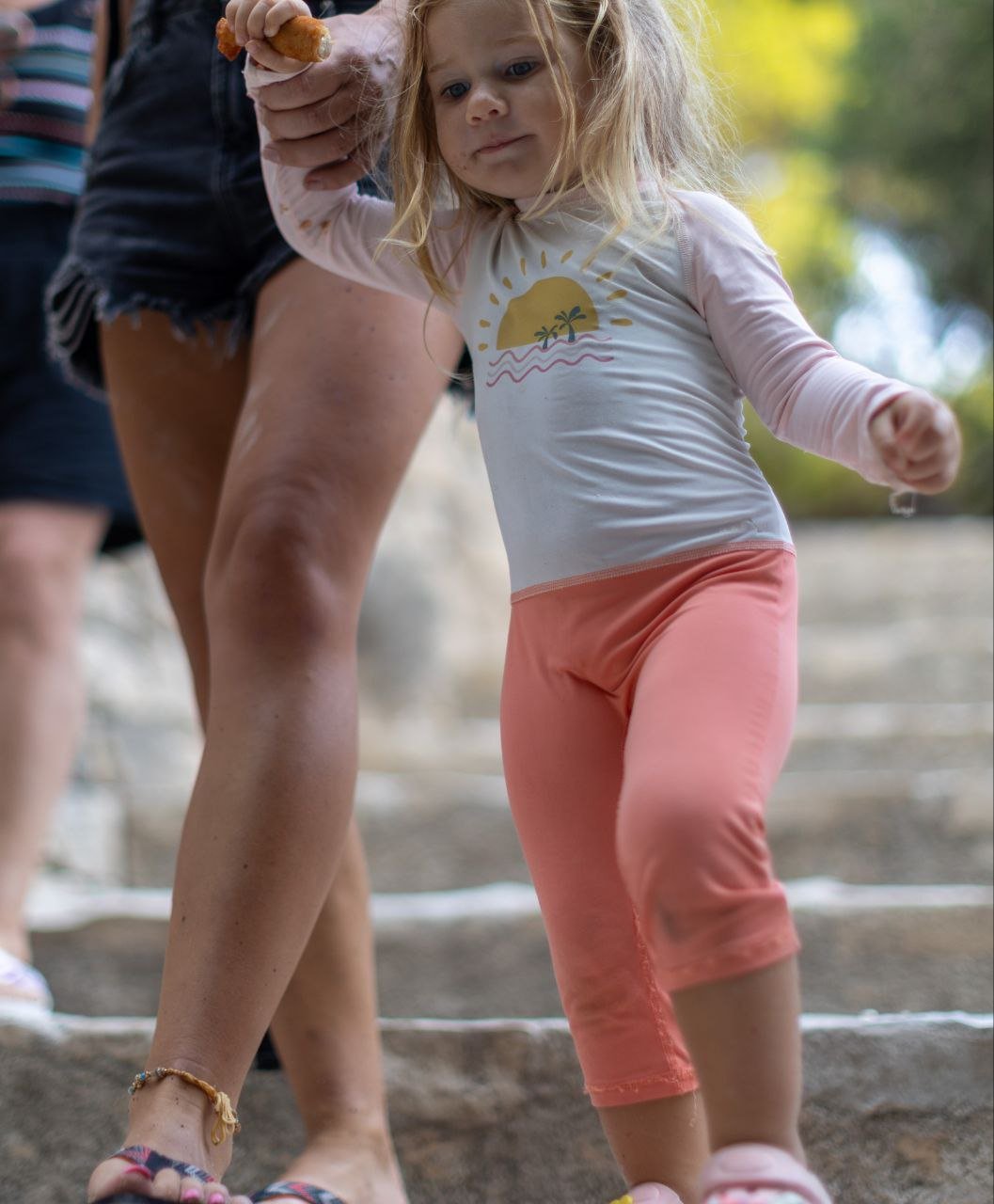 A young girl with blonde hair in a ponytail holding hands with an adult, walking outdoors on stone steps. The girl is wearing a white long sleeve shirt with a sunset and palm tree graphic and pink pants.