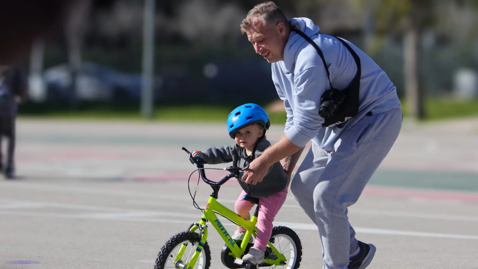 An adult helping a young child learn to ride a bicycle outdoors on a sunny day, both wearing helmets.
