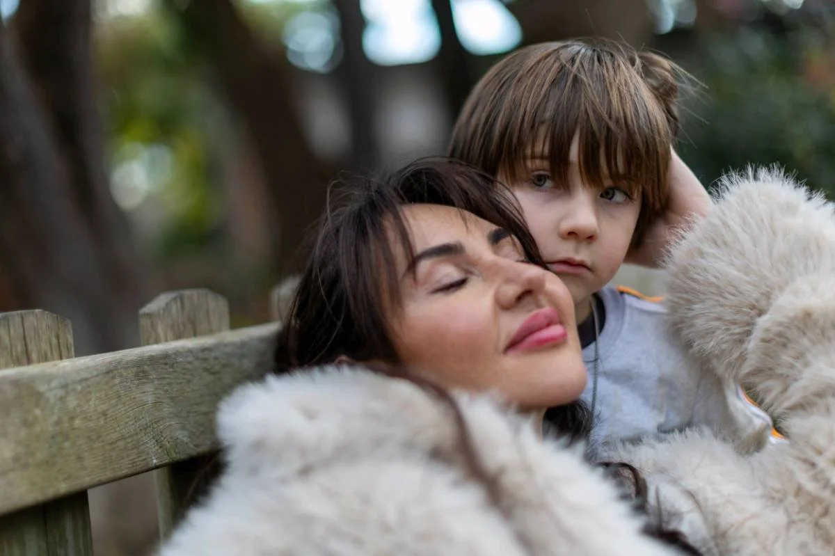 A woman and a young boy sitting close together on a wooden bench outdoors, with the woman resting her head on the woman's shoulder. They have relaxed expressions, and a fluffy white dog is in front of them. The background is blurred greenery and tree