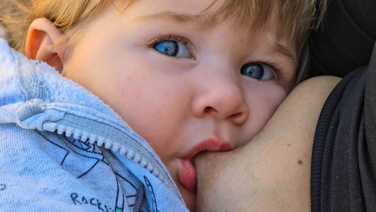Close-up of a young child with blue eyes resting on their mother's shoulder, suckling on her breast, wearing a light blue hoodie with a zipper.
