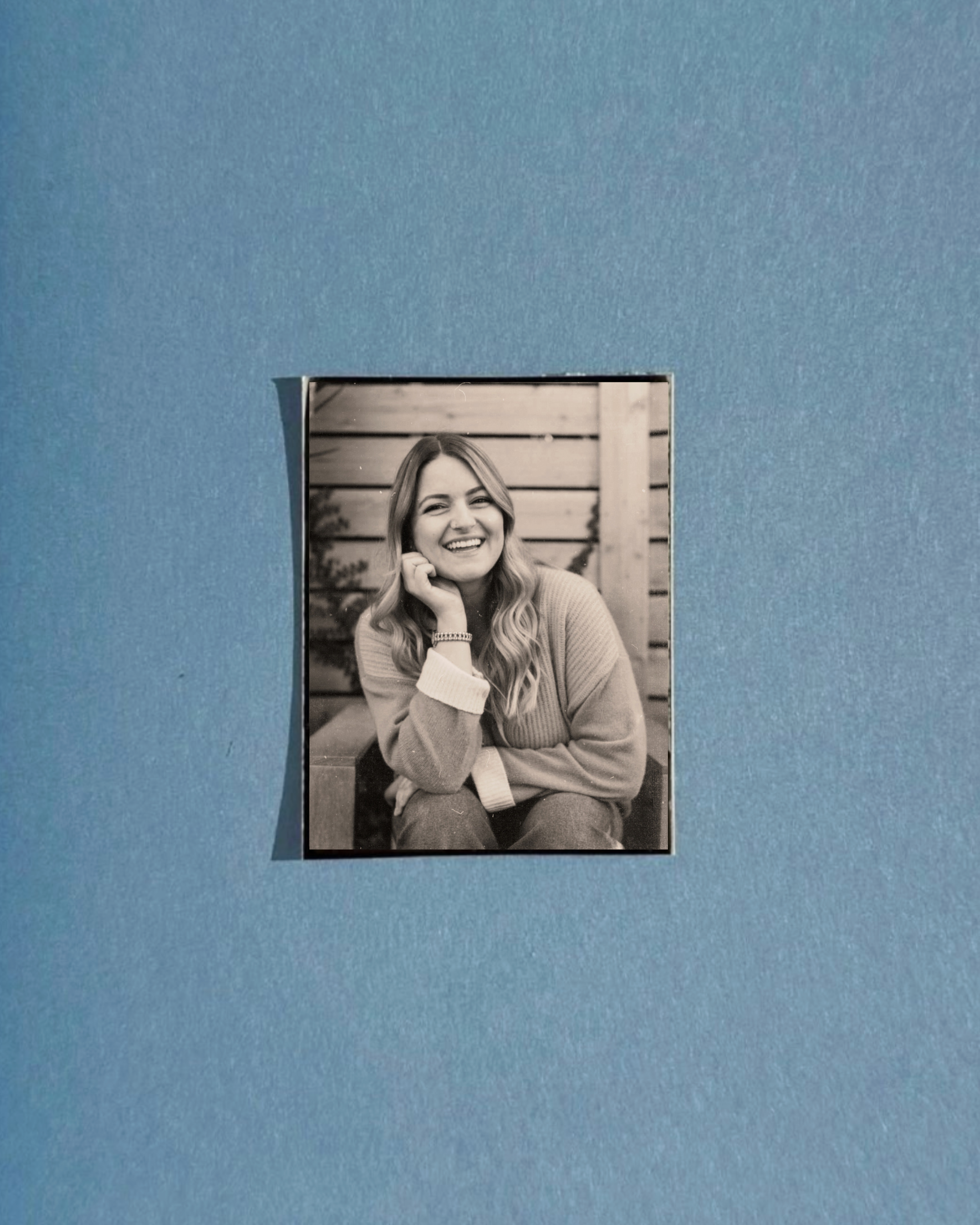 Black and white photograph of a woman with long wavy hair, smiling and sitting in front of a wooden fence, placed in the center of a blue background.