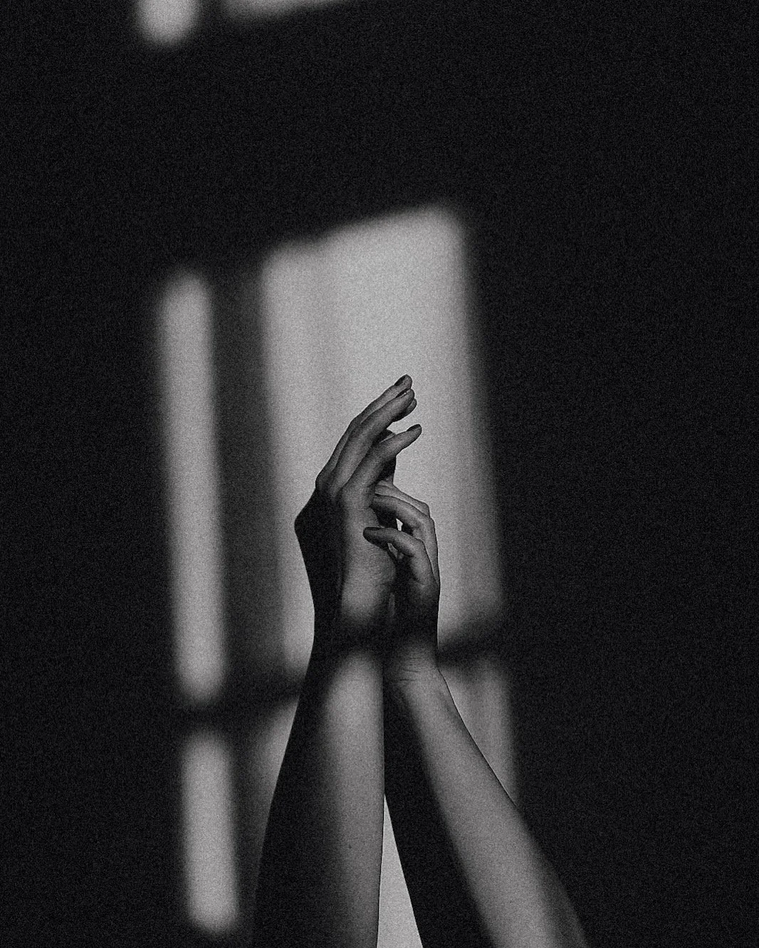 Black and white photograph of a person's hands clasped together, with soft light creating shadows on the arms and hands, against a dark background.