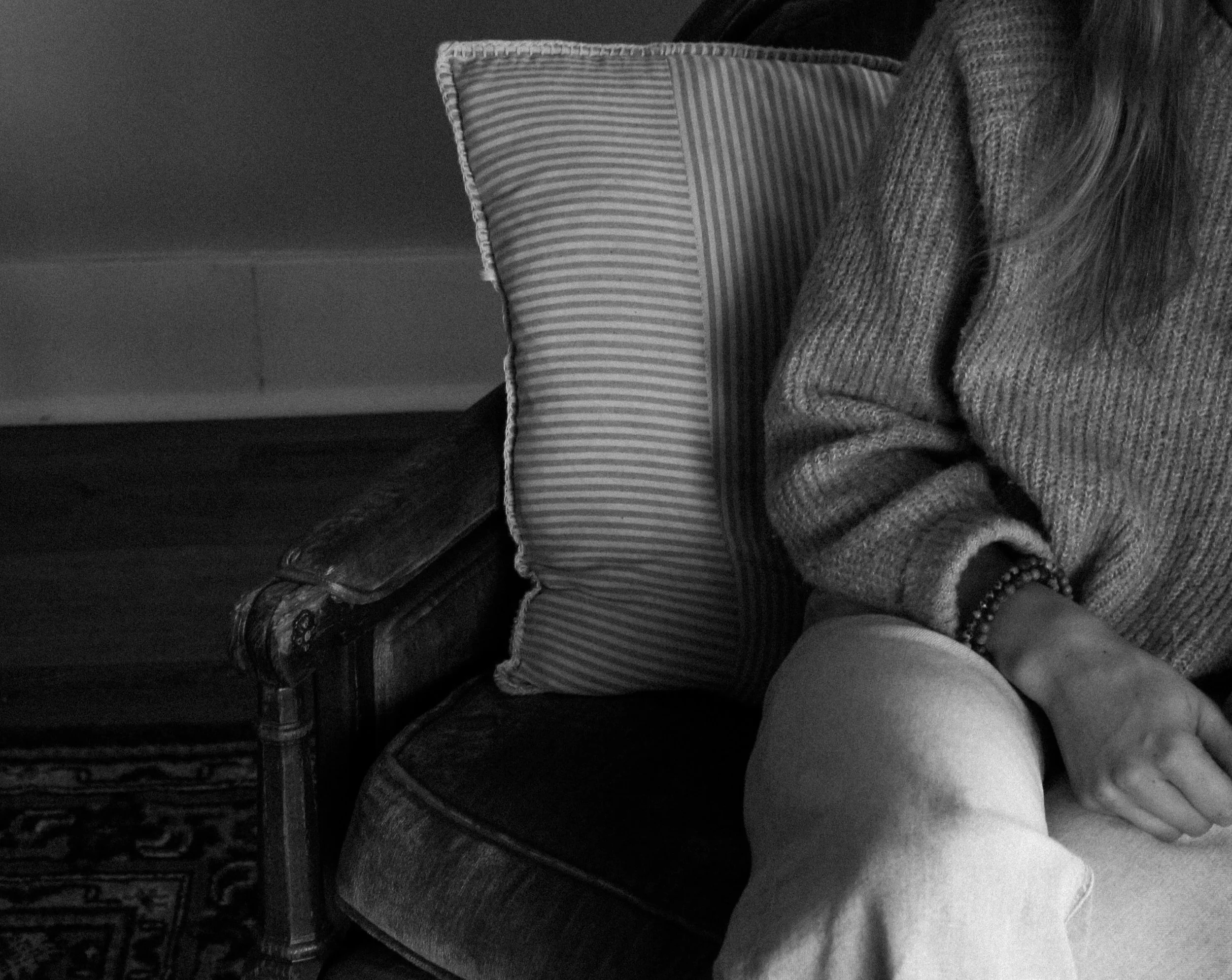Partial view of a person sitting on a vintage sofa with a striped pillow, wearing a sweater, and a bracelet on their wrist.