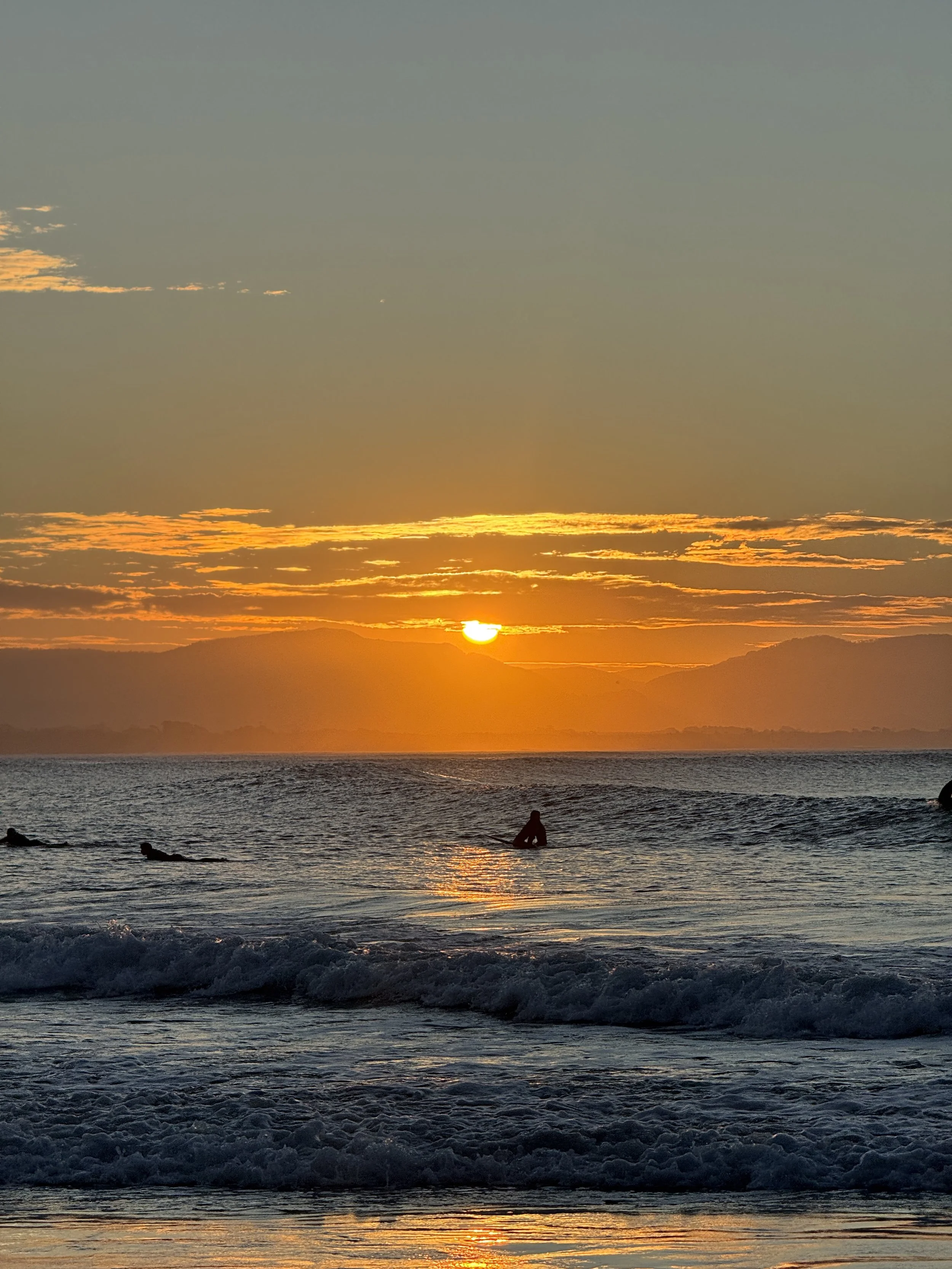 Sunset over the ocean with a few surfers in the water and mountains in the background.
