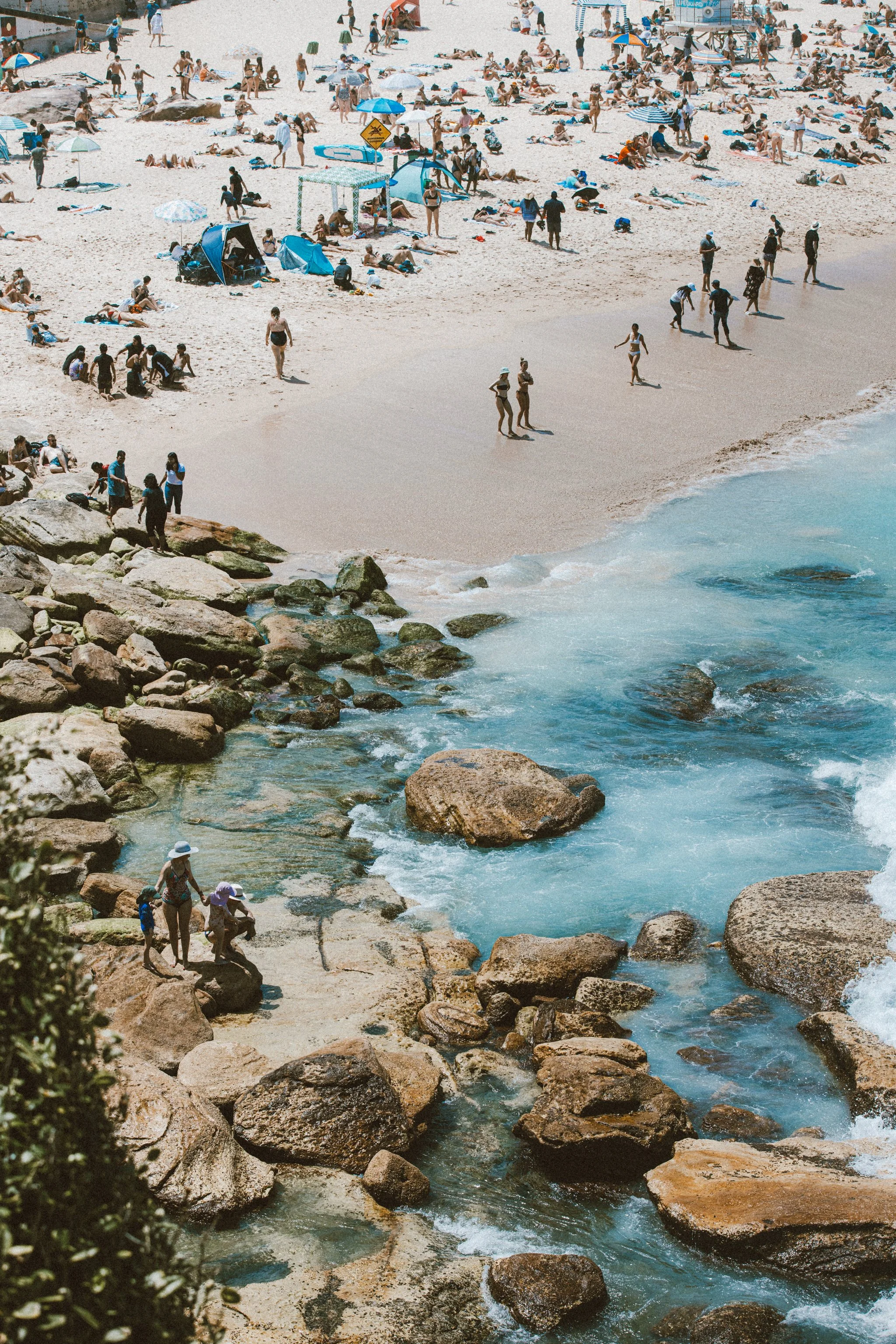 A crowded beach with many people sunbathing, walking, and playing near the shore. There are umbrellas, tents, and beach equipment scattered across the sand. In the foreground, a rocky shoreline with some individuals exploring near the water, which is blue with waves crashing against the rocks.