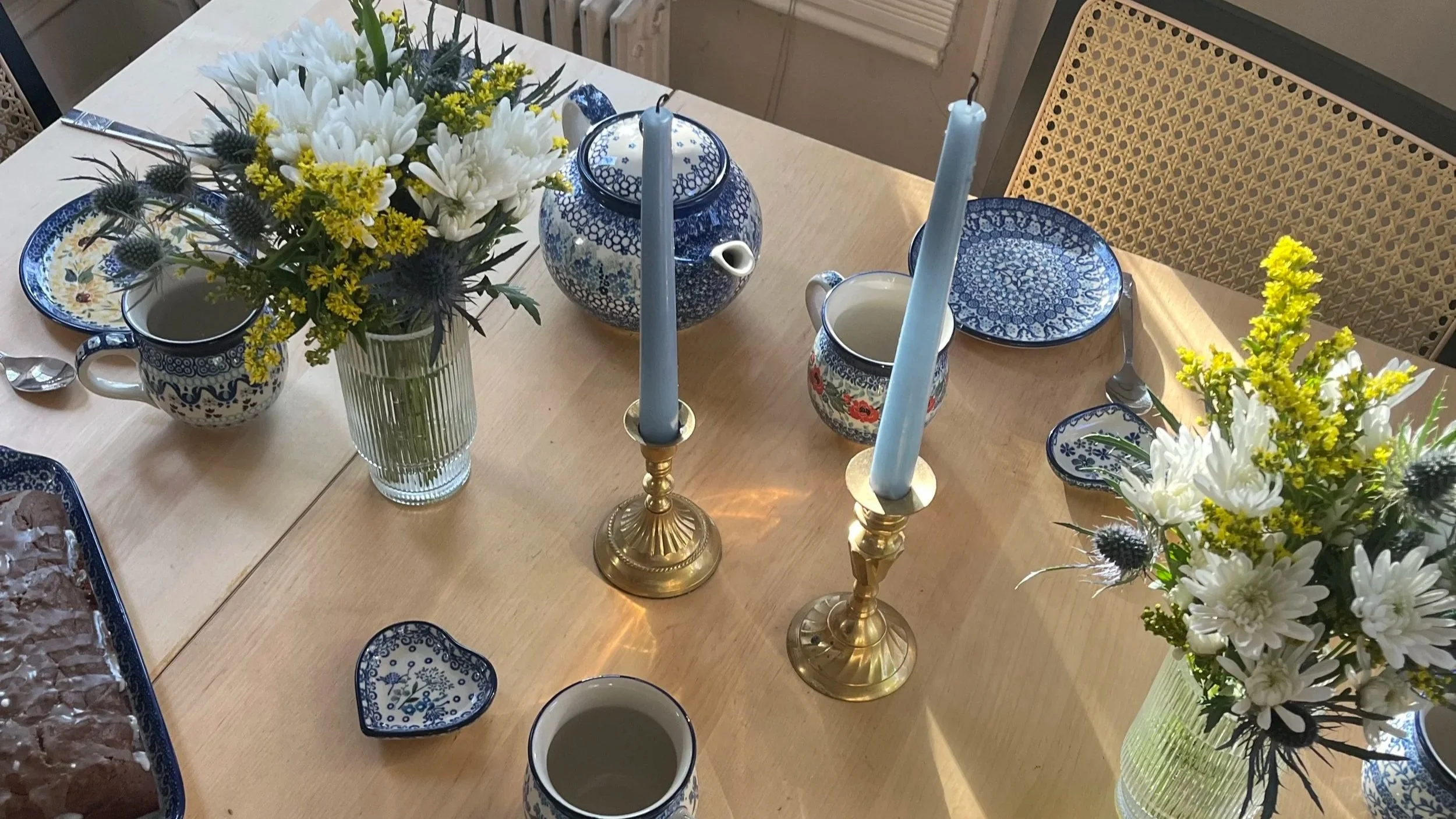 A dining table set with blue and white patterned plates, mugs, and a teapot. Two vases with white daisies, yellow flowers, and greenery. Two brass candlesticks with tall blue candles. The table has a natural wood finish, and there are chairs with woven backs.