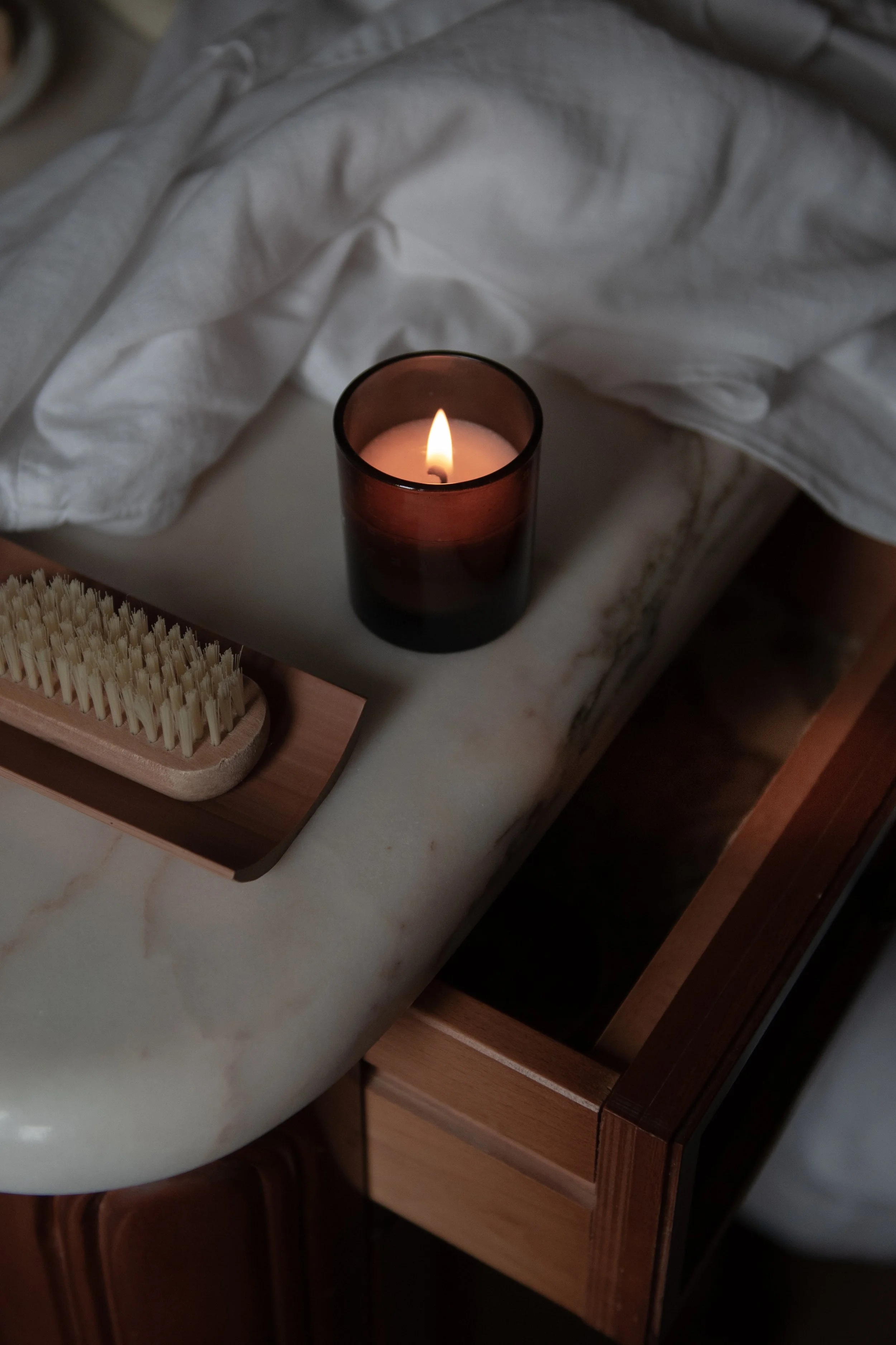A lit candle in a dark glass holder on a marble bedside table with a wooden brush and a wooden drawer.