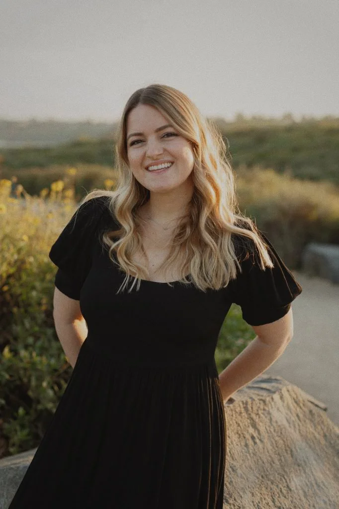 A young woman with blonde wavy hair smiling outdoors during golden hour, wearing a black dress with puff sleeves, standing near a rock and greenery.
