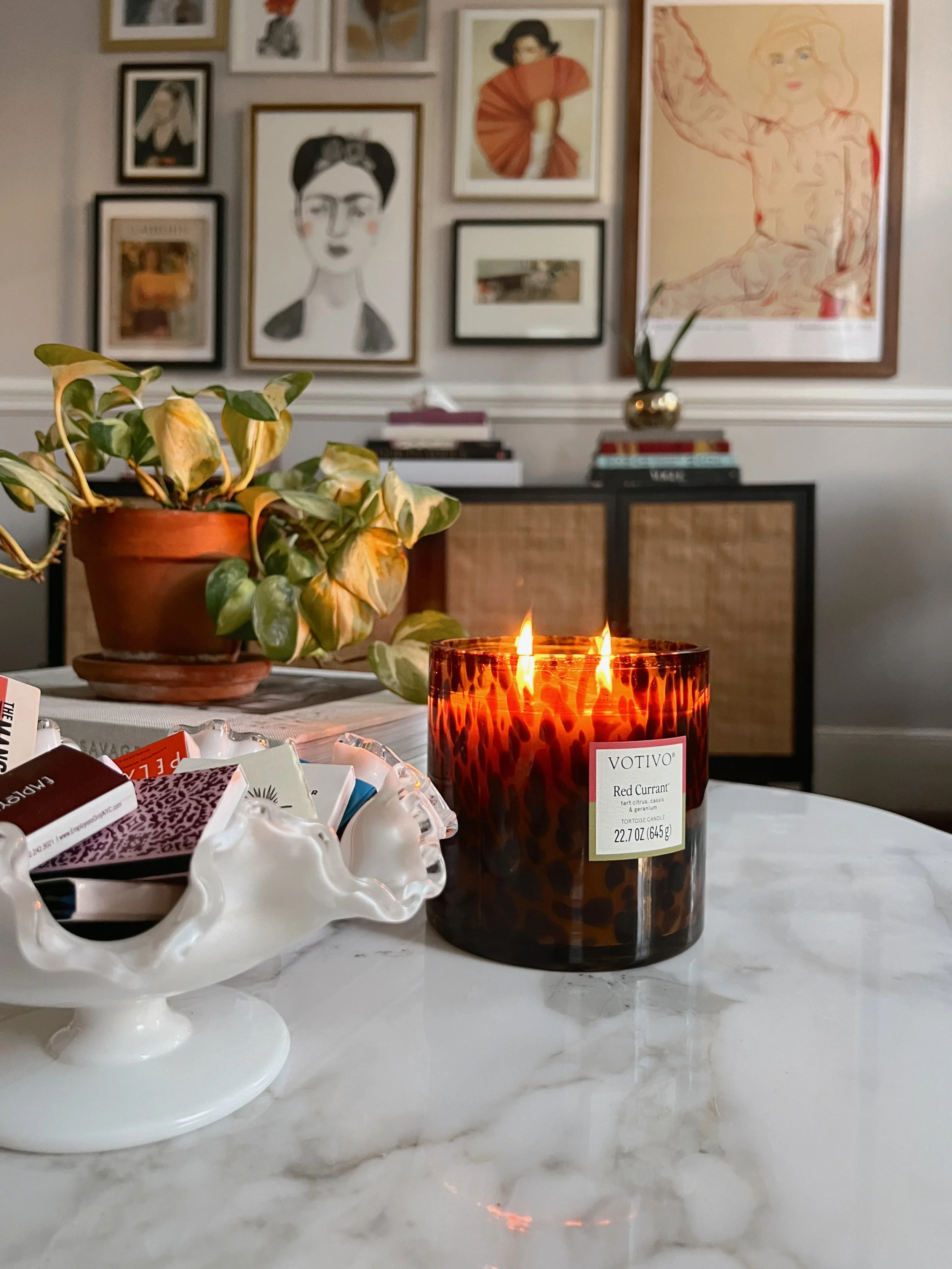 A lit tortoise shell scented candle labeled 'Votivo Red Currant' sits on a marble table next to a white ceramic dish filled with assorted small items, with a potted plant with variegated leaves in the background, and a gallery wall with framed artwork hanging in a living room.