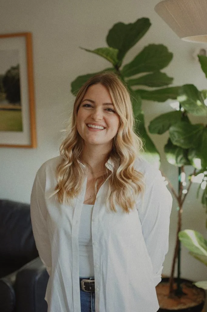 A young woman with blonde, wavy hair wearing a white shirt and smiling indoors with green plants and framed pictures in the background.