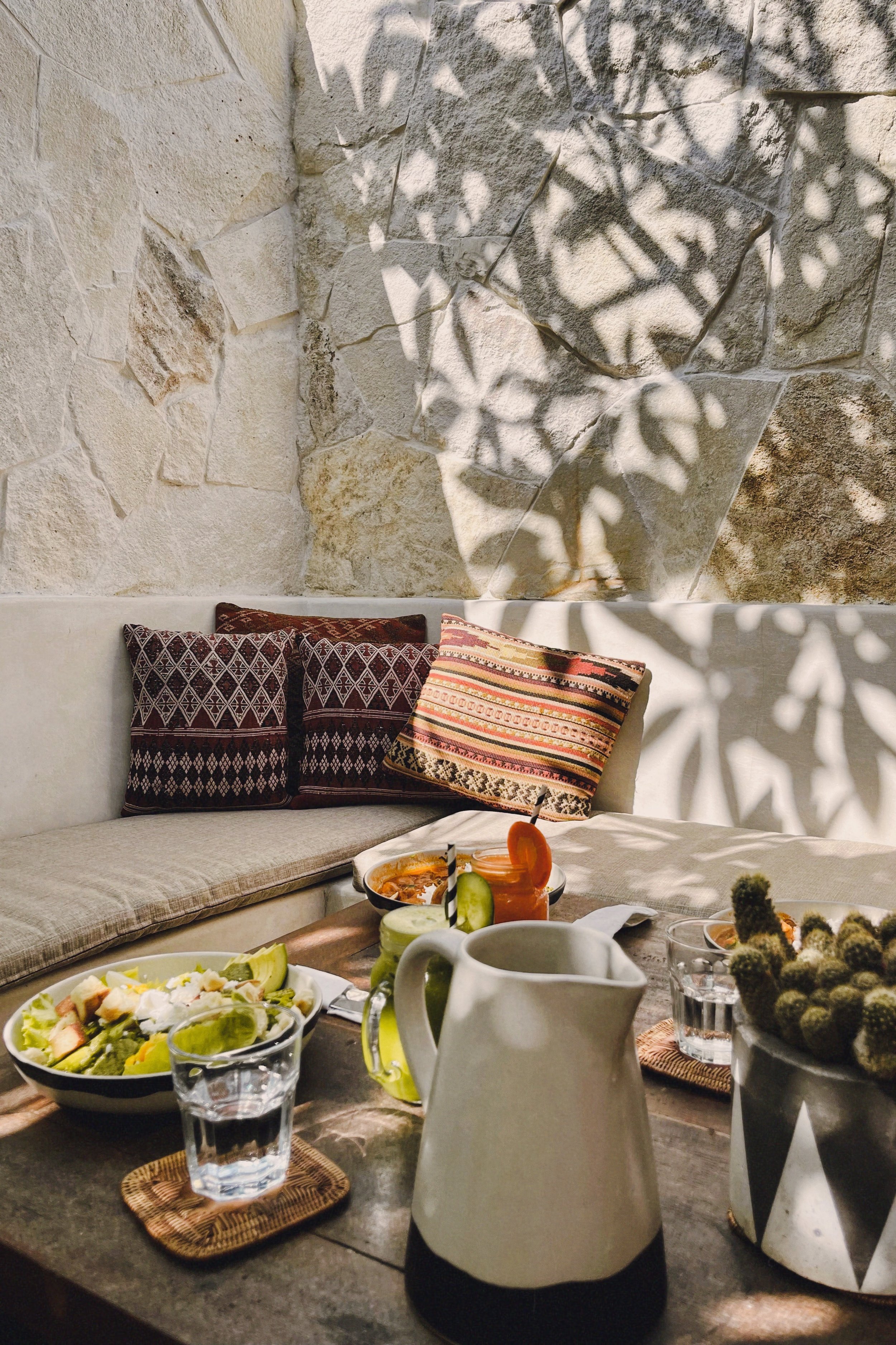 A cozy indoor dining area with a stone wall, patterned pillows, and a table set with salads, drinks, and small plates, illuminated by sunlight and shadows of leaves.