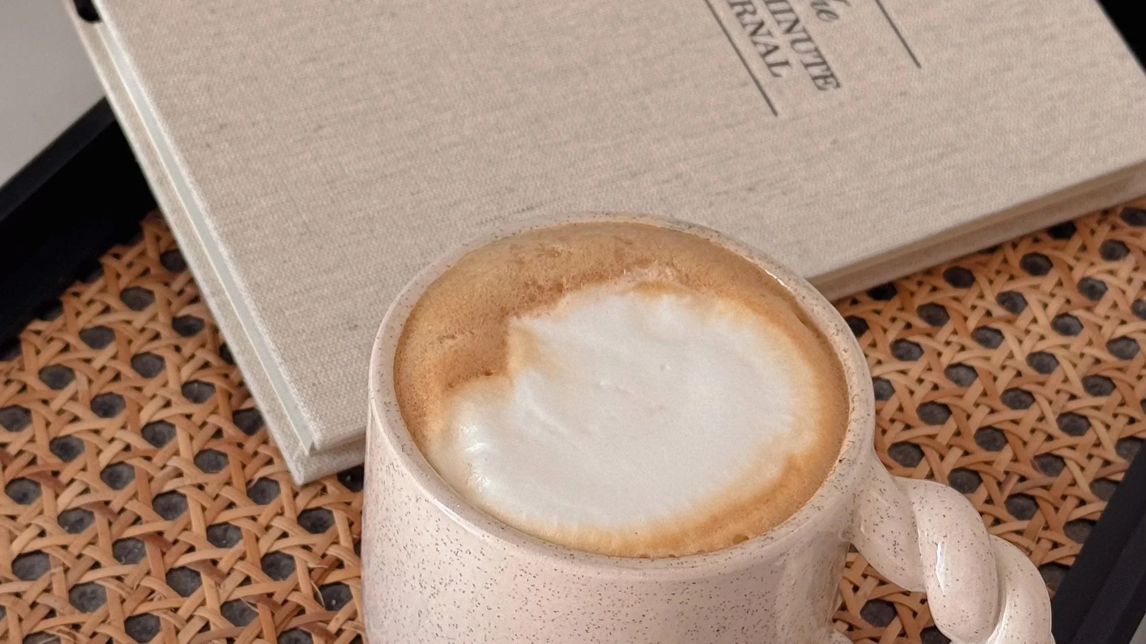 A white ceramic coffee mug filled with a frothy cappuccino resting on a woven rattan surface, with a closed beige book nearby.