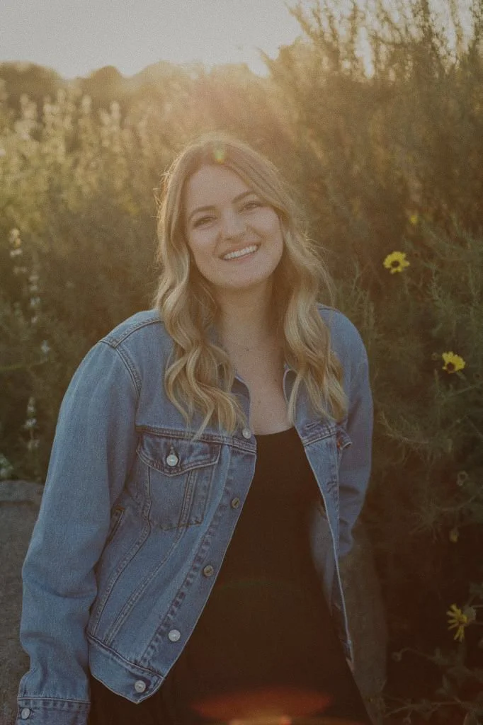 A young woman with long blonde hair smiling outdoors at sunset, wearing a denim jacket over a black top.