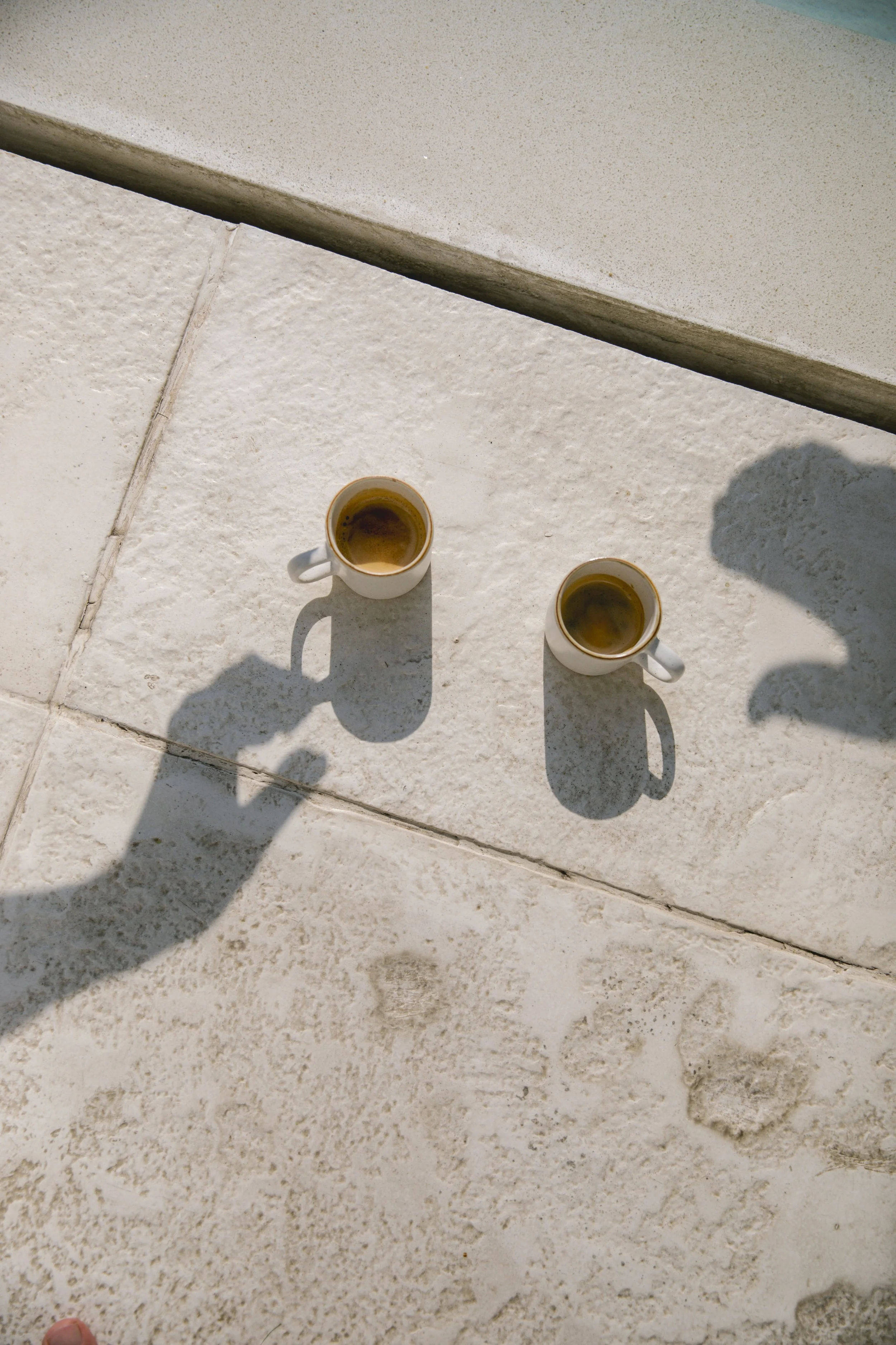 Two cups of espresso on a tiled outdoor surface, casting long shadows in sunlight.