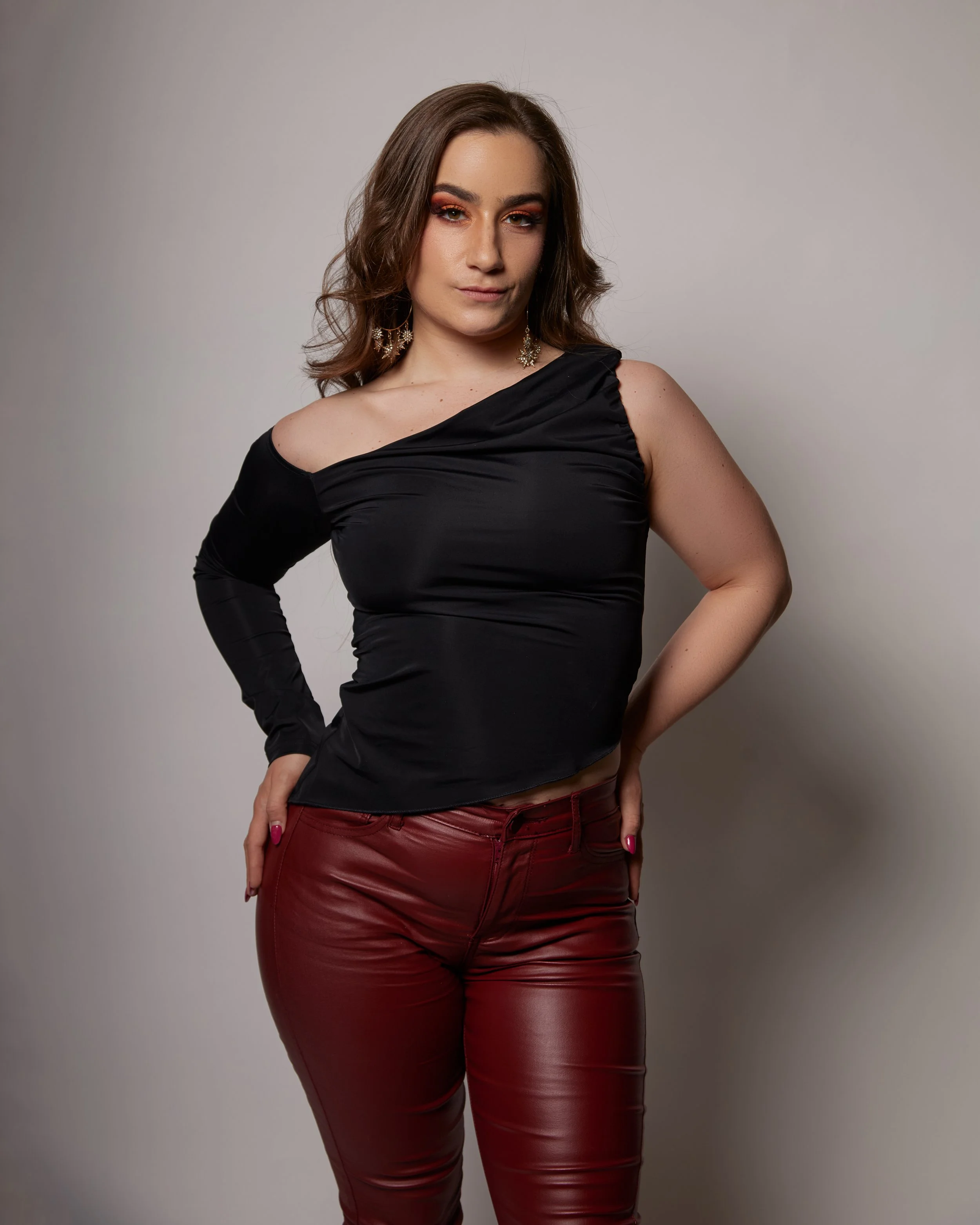 Cat Rojo wearing large sparkly hoop earrings, posed with her hands on her hips. She is wearing a black top with read pleather pants for a recent photoshoot.