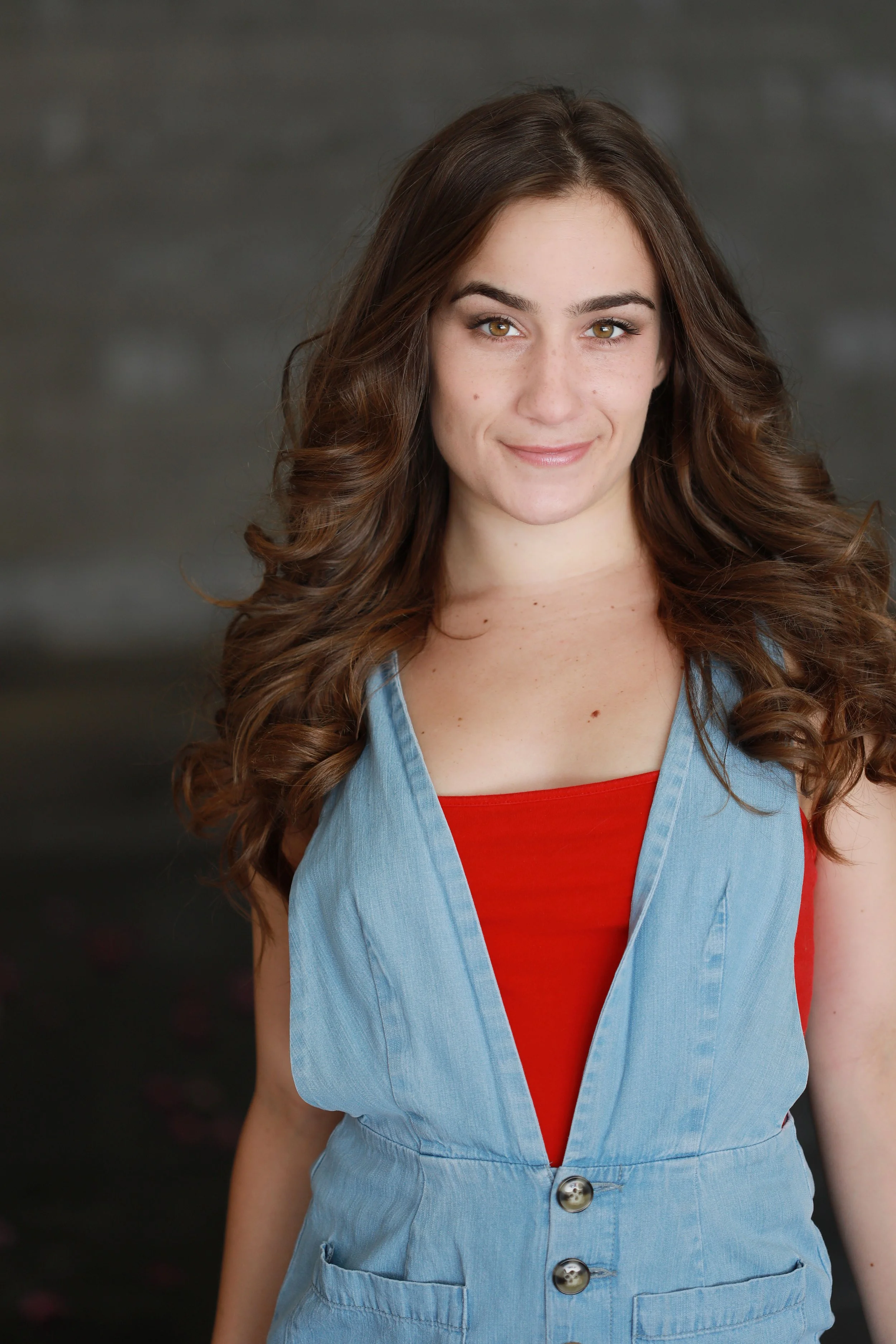 Cat Rojo 3/4 headshot wearing a red top and jean overalls on top, smiling and looking into the camera