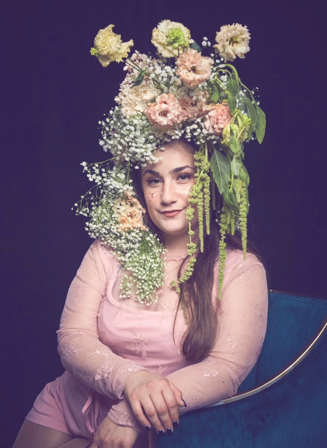 Cat Rojo seated in a blue chair looking straight at the camera wearing an elaborate floral headpiece with draping greenery at the Solstice Soirée for The Old Globe in San Diego