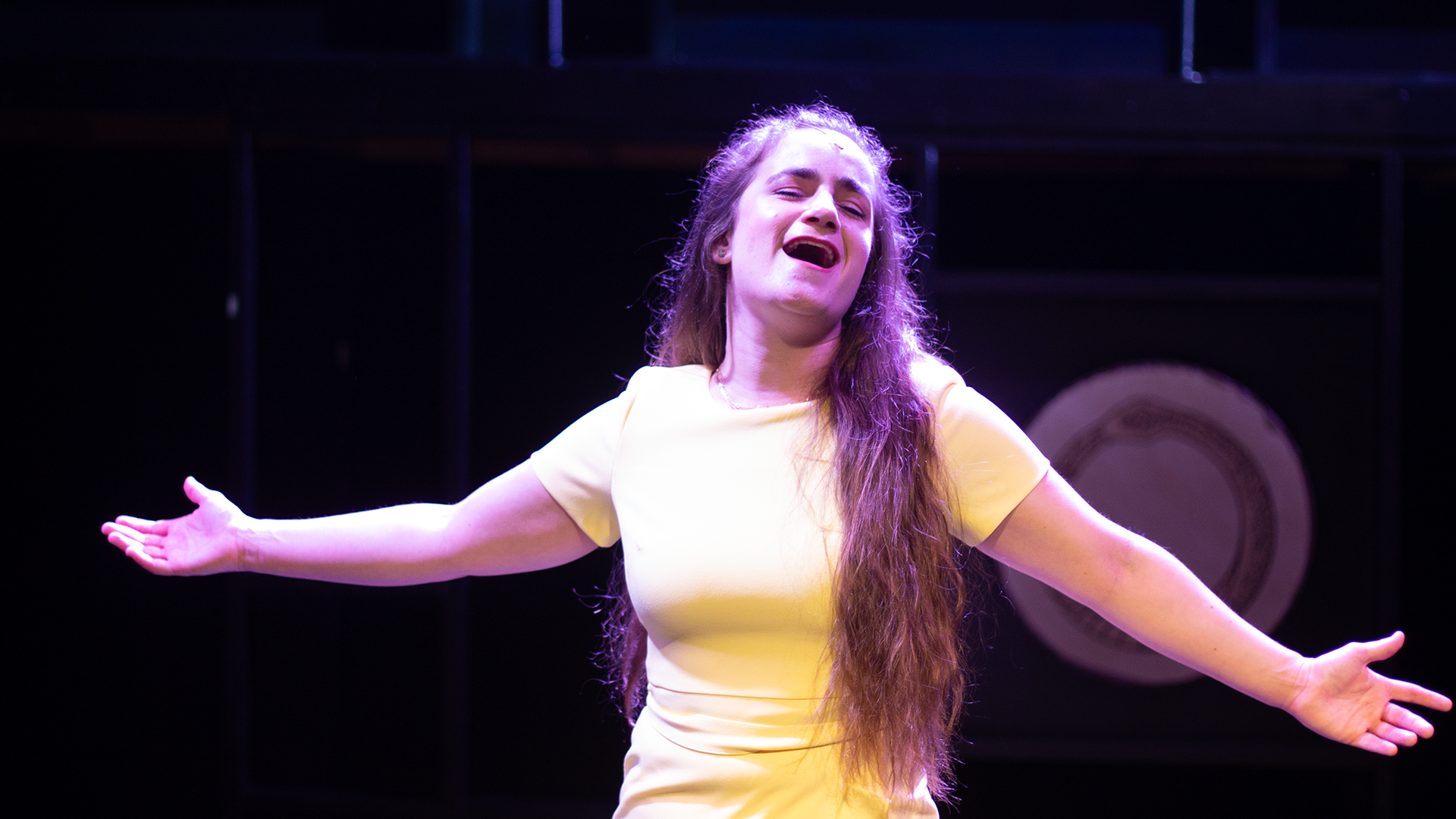 Cat Rojo singing soulfully with her arms open in a bright yellow audition dress during "Climbing Uphill" in The Last Five Years the Musical