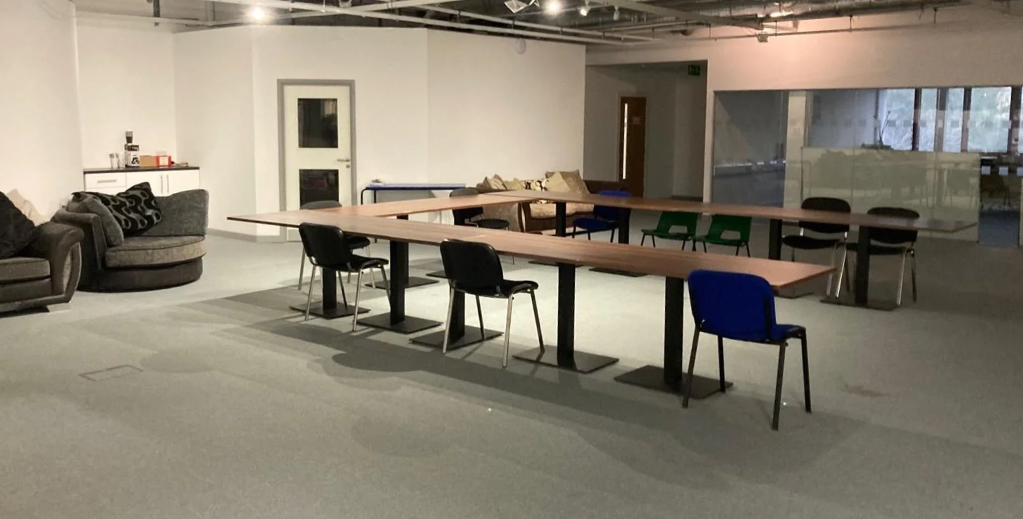 An empty modern office or conference room with long wooden tables, black and colored chairs, a gray sofa with pillows, and a white cabinet with coffee supplies. Large windows on the right side with a glass wall, and a door in the background.