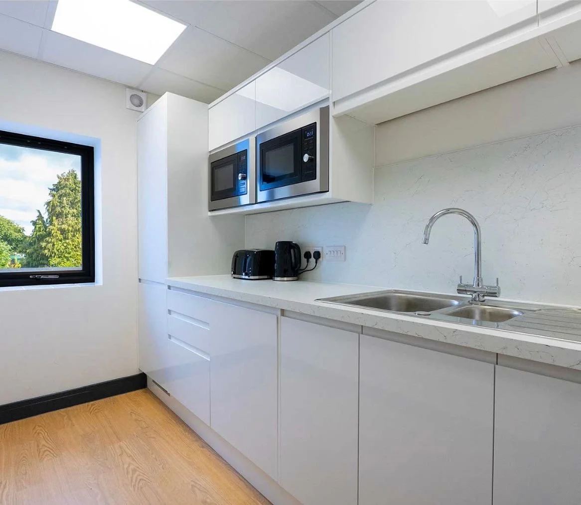 Modern kitchen with white cabinets, microwave, toaster, kettle, stainless steel sink, window showing trees outside, and wooden flooring.