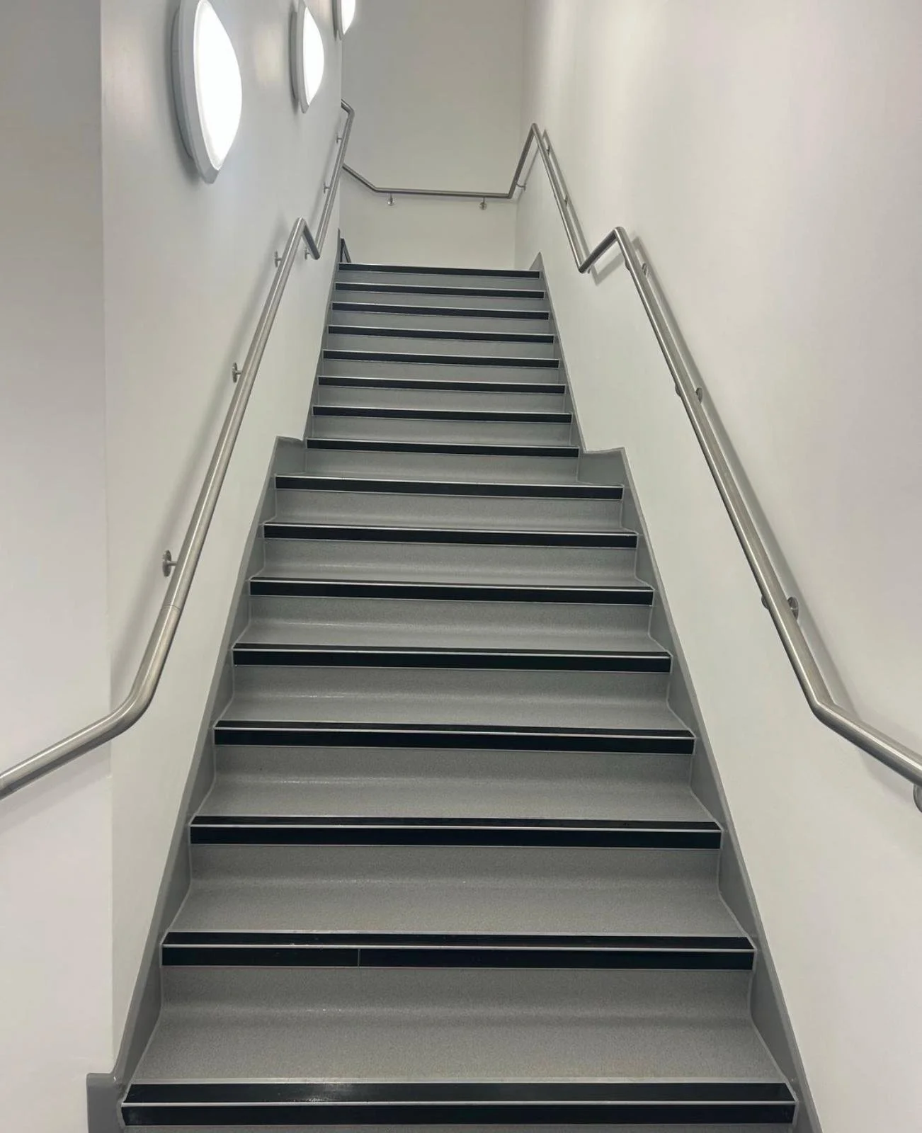 A clean indoor staircase with gray steps, black anti-slip strips, metal handrails on both sides, white walls, and modern circular wall lights.