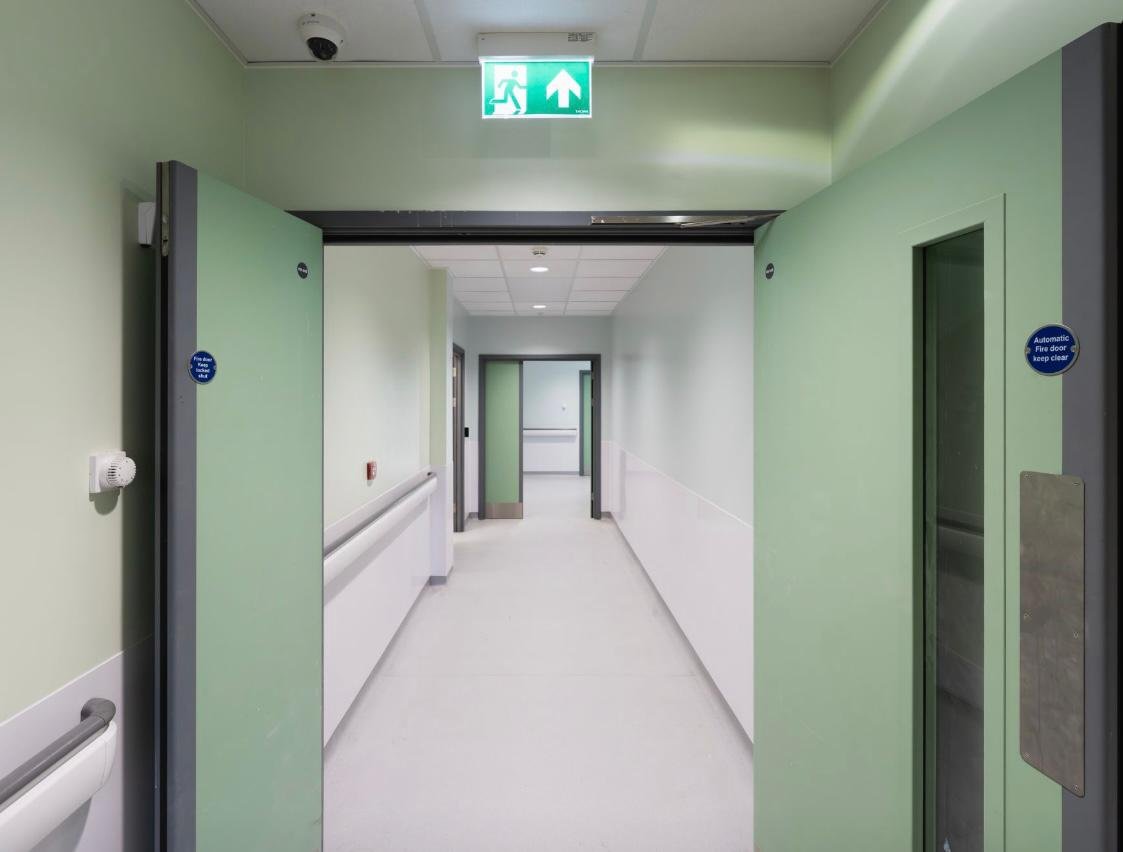 Hospital corridor with automatic sliding doors at the entrance and an exit sign with a running figure and arrow above.