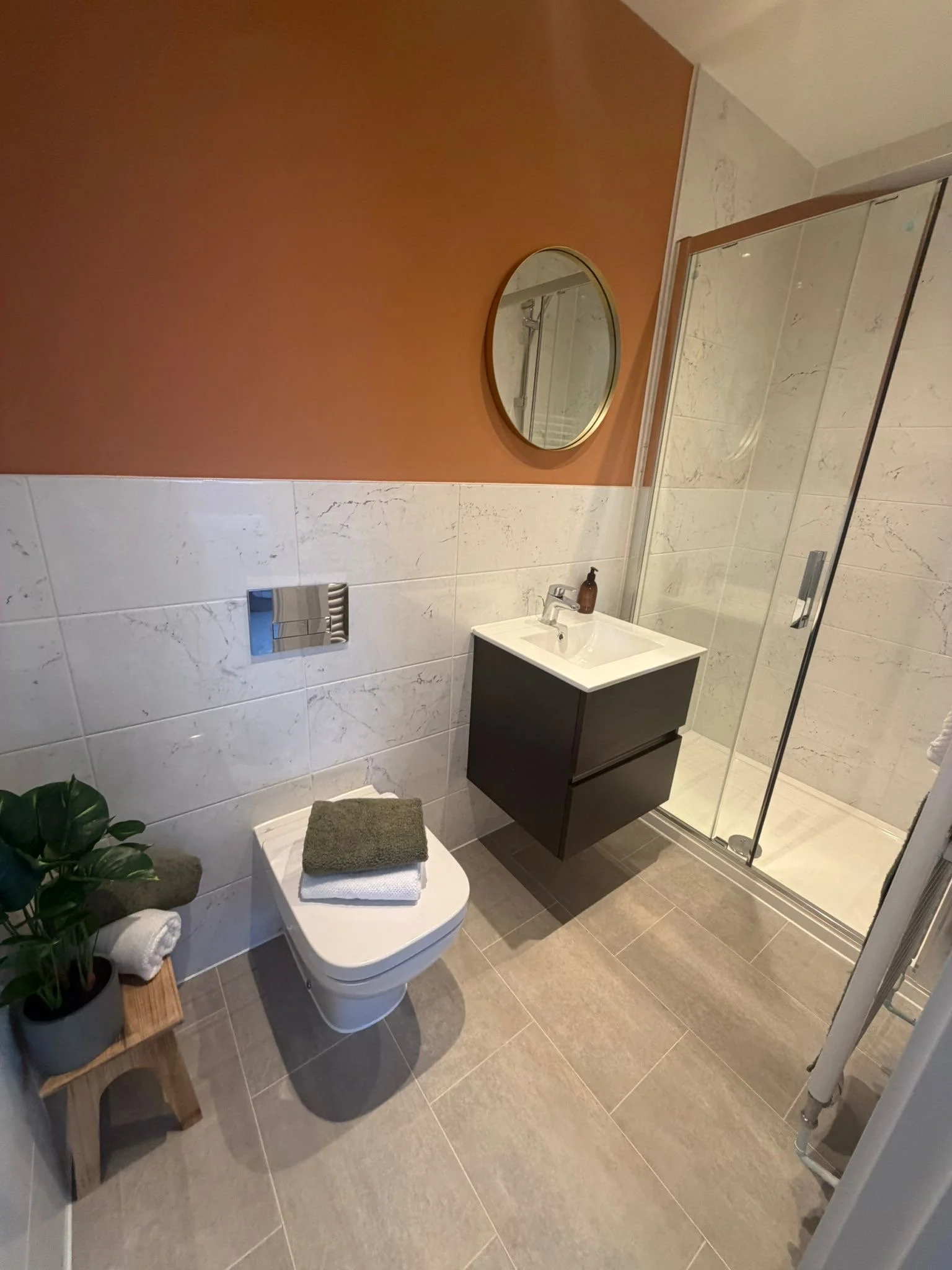 Modern bathroom with beige tiles, a white toilet with folded towels on top, a small black vanity with a white sink, a round mirror, a glass shower enclosure, and a potted plant near the toilet.