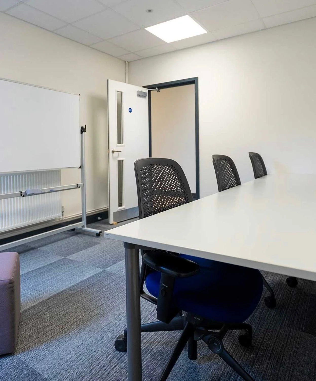 Empty conference room with white table, black mesh chairs, whiteboard, closed door, and gray carpet.