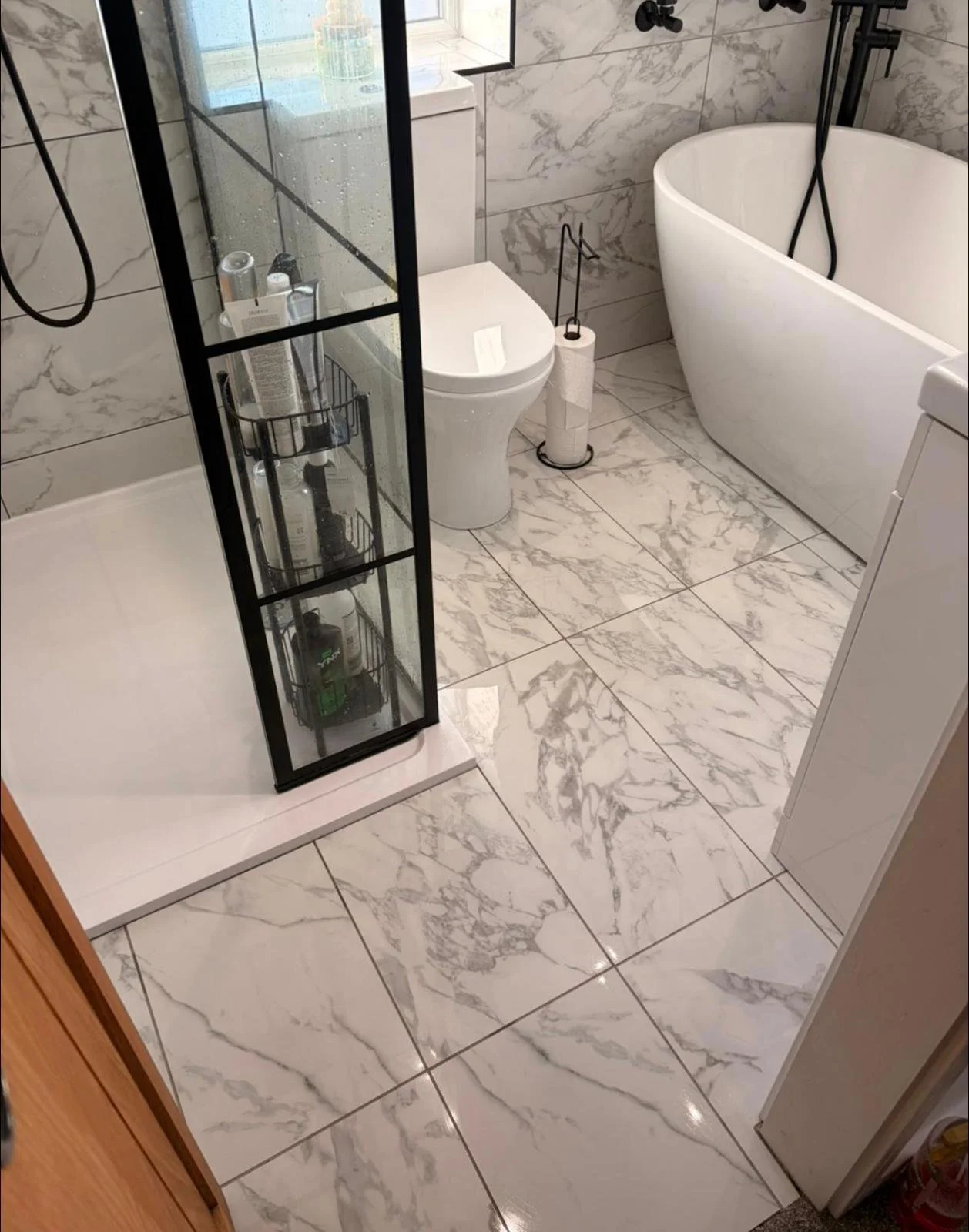 Modern bathroom with marble floor and walls, featuring a shower with a glass door, a white toilet, a free-standing bathtub, and a washing machine.