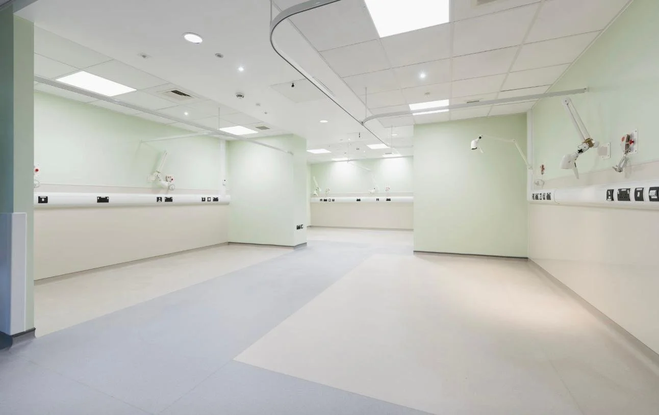 Empty hospital or medical room with white walls, ceiling, and light-colored flooring, featuring medical equipment and outlets along the walls.